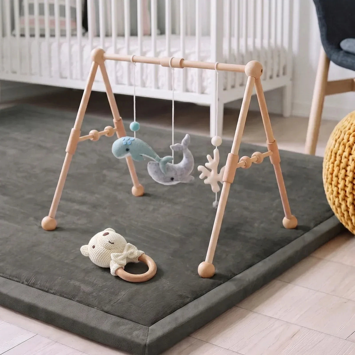 L'arche d'éveil en bois posée sur le tapis d'éveil gris, cinq jouets en feutrine suspendus, le hochet crochet posé devant, dans une chambre d'enfant